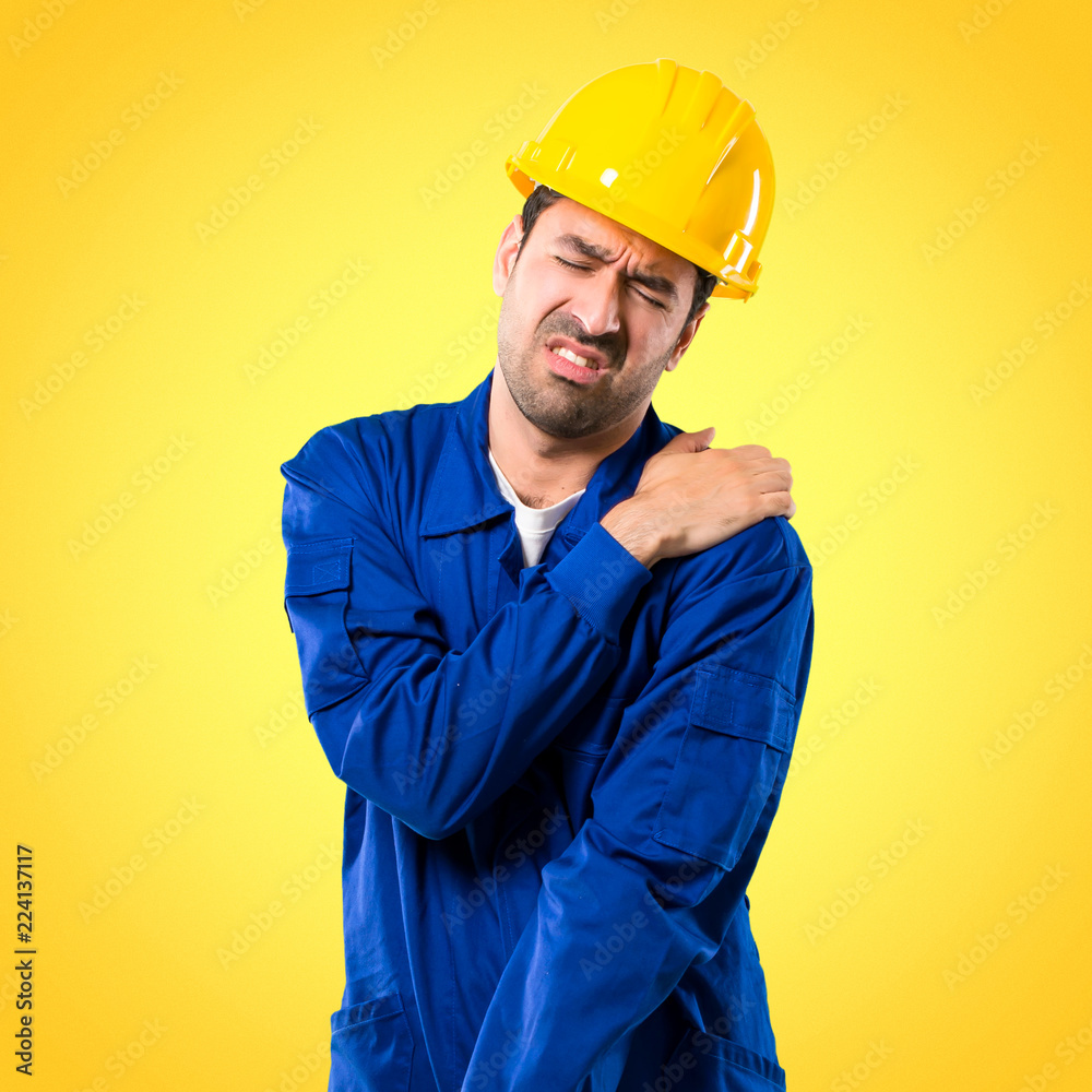 Young workman with helmet suffering from pain in shoulder for having made an effort on yellow background