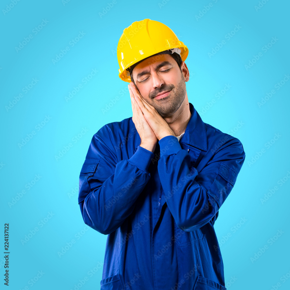 Young workman with helmet making sleep gesture. Adorable and sweet expression on blue background Young workman with helmet making sleep gesture. Adorable and sweet expression on blue background