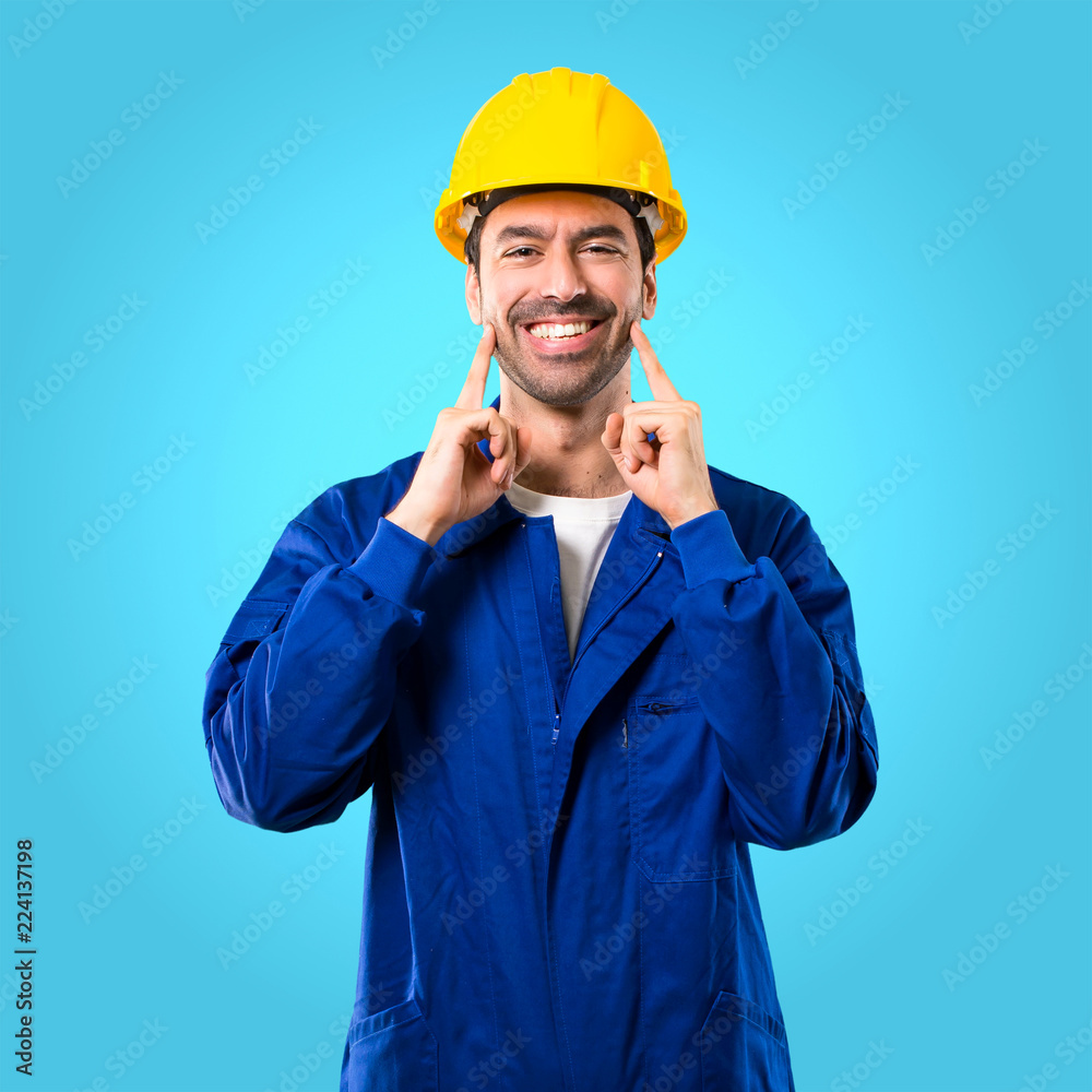 Young workman with helmet smiling with a happy and pleasant expression while pointing mouth and face with fingers on blue background