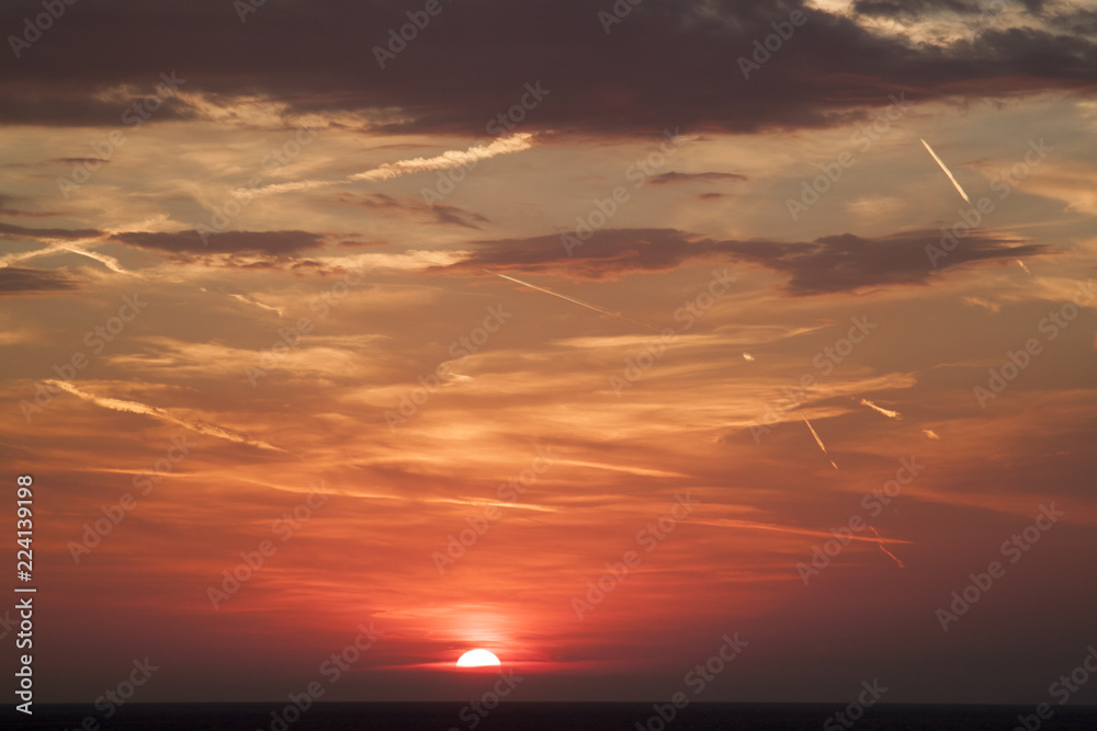 beautiful view of the sun hiding behind the sea surface.  the sunset sky is orange with clouds and condensation traces