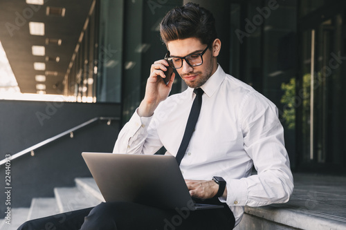 Portrait of serious businessman dressed in formal suit sitting outside glass building with laptop, and speaking on cell phone