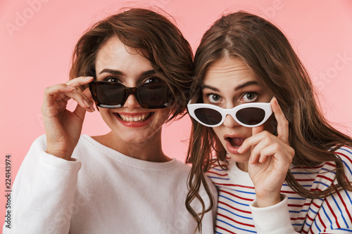 Emotional young women friends isolated over pink wall background wearing sunglasses.