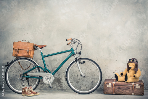 Retro bicycle with leather mailman's bag, old sneakers and Teddy Bear toy in leather aviator's hat and goggles sitting on aged classic travel suitcase front concrete wall. Vintage style filtered photo