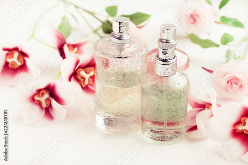 Light aroma perfume bottles wet with water drops surrounded by pink flowers on white bathroom shelf. Pastel colors toned. Feminine delicate scents product.
