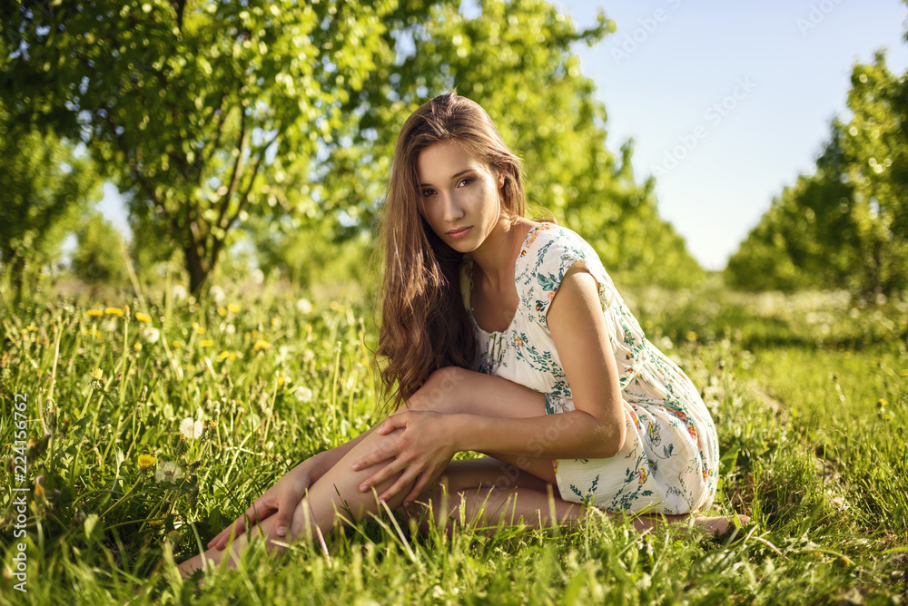 A brown-haired girl sitting leisurely on grass in a picturesque orchard.