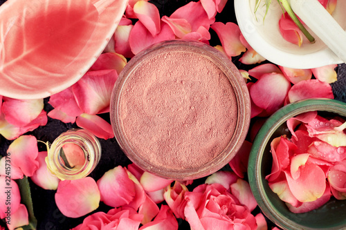 Pink cosmetic clay closeup with rose petals, ceramic bowls on black table, homemade skincare mask preparation