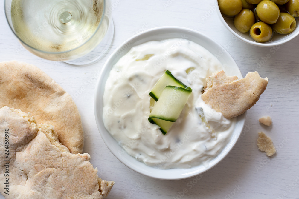 Tzatziki in white ceramic bowl with a cucumber slice and a piece of pita bread next to pita bread, olives and a glass of white wine from above.