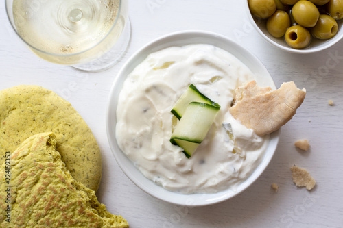 Tzatziki in white ceramic bowl with a cucumber slice and a piece of pita bread next to garlic and coriander pita bread, olives and a glass of white wine from above.