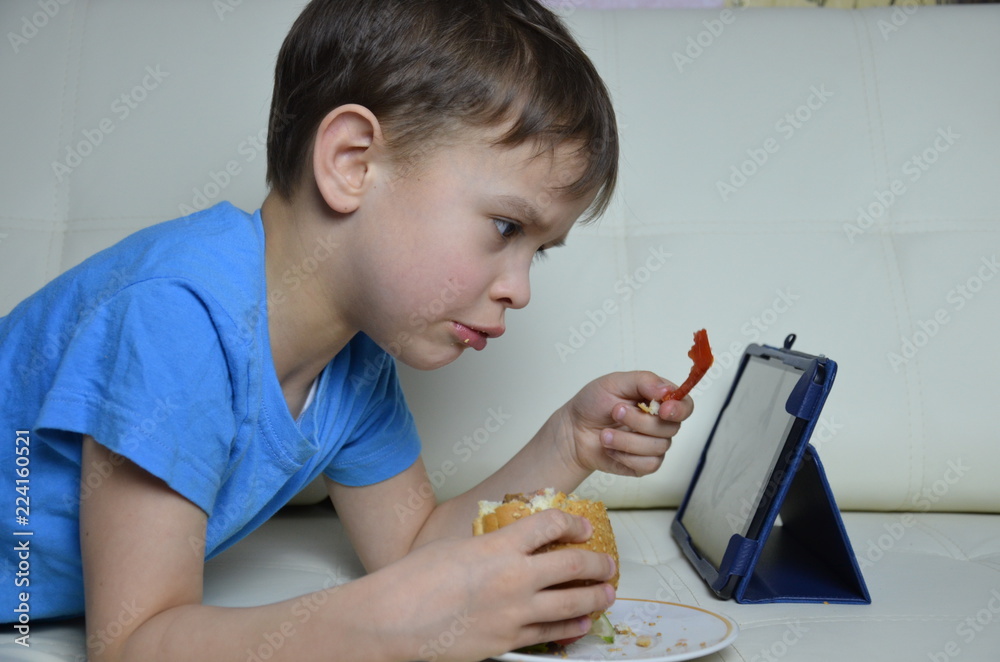 cute boy at home on the couch, eating a hamburger and looking at the tablet, watching cartoons or talking with friends he eat burgers and look on the tablet. he focused on displayed on the screen. cute boy at home on the couch, eating a hamburger and looking at the tablet, watching cartoons or talking with friends he eat burgers and look on the tablet. he focused on displayed on the screen.