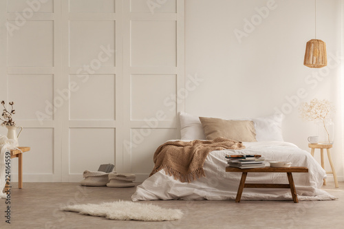 Fur and wooden stool in front of bed with blanket in white bedroom interior with lamp. Real photo photo