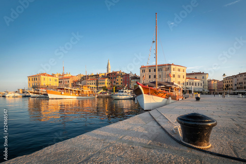 Fototapeta Naklejka Na Ścianę i Meble -  Motorboats and boats on water in port of Rovinj. Medieval vintage houses of old town. Yachts landing, high tower of Church of Saint Euphemia. Morning sunrise blue sky