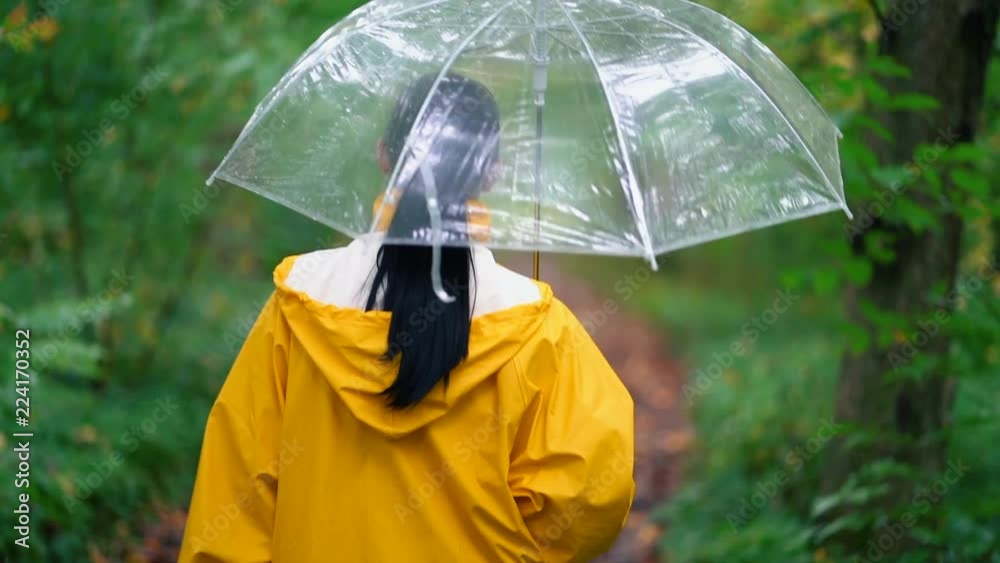 Young pretty woman in yellow raincoat walking in autumn park or forest alone. Girl with transparent umbrella. Rainy weather, nature background.