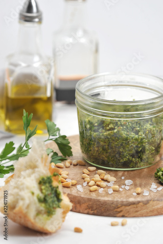 Green pesto sauce in a jar, large salt, garlic clove, pine nuts on a wooden plate. In the frame, olive oil, a slice of ciabatta. Close-up. Macro photography. Vertical orientation of the frame.