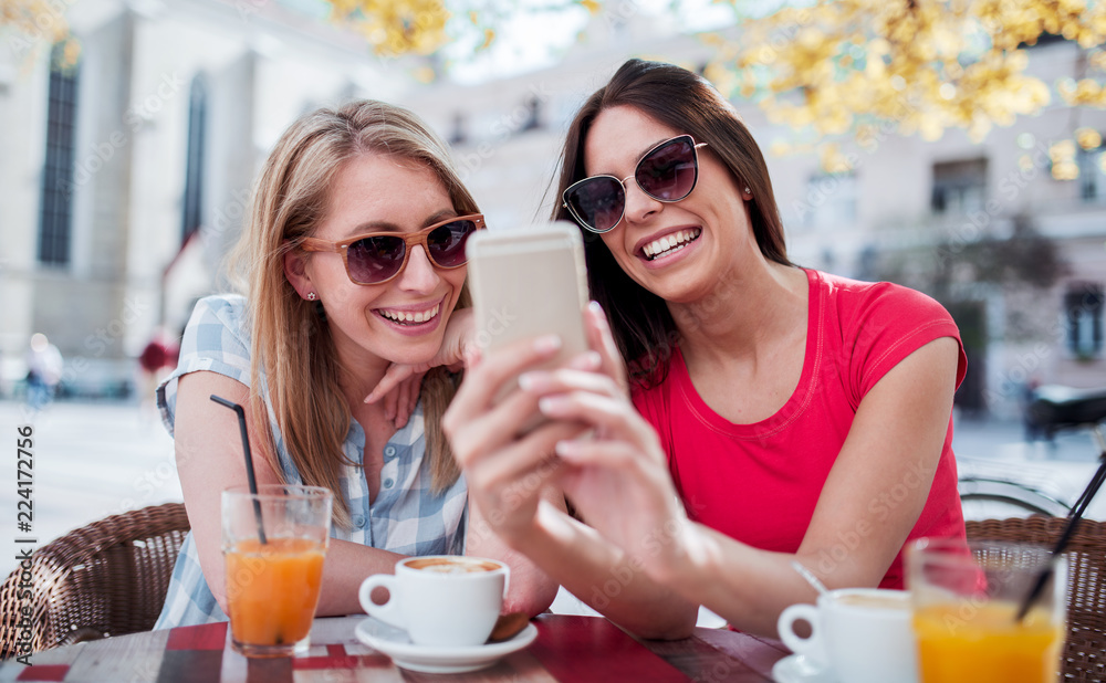 Friends meeting in a cafe. Young women drinking coffee and have fun with mobile phone. Consumerism, lifestyle concept Friends meeting in a cafe. Young women drinking coffee and have fun with mobile phone. Consumerism, lifestyle concept