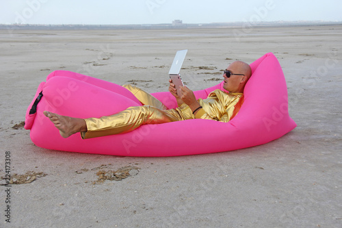 Frilanser freak. A young handsome bald man in gold metal clothes and a suit is lying on a pink inflatable sofa chair and working at the computer, laptop photo