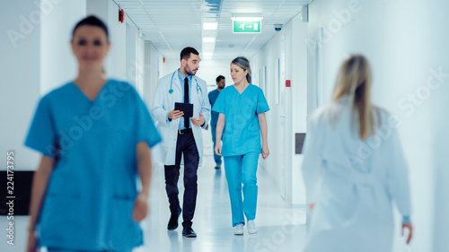 Surgeon and Female Doctor Walk Through Hospital Hallway, They Consult Digital Tablet Computer while Talking about Patient's Health. Modern Bright Hospital with Professional Staff.