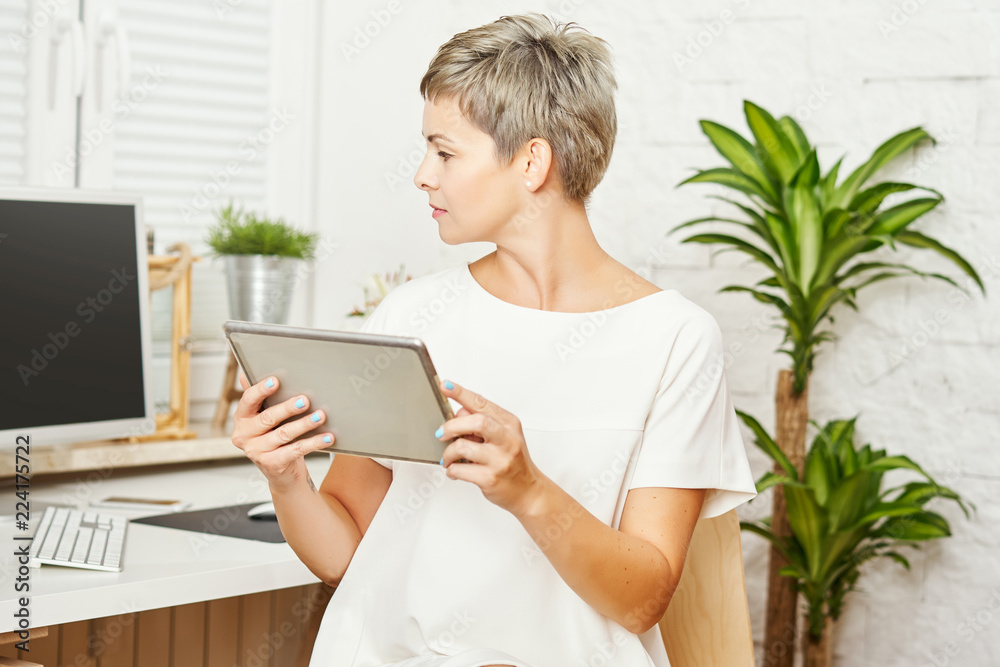 beautiful business woman in a white dress sitting at a desk and works on a tablet in a home office beautiful business woman in a white dress sitting at a desk and works on a tablet in a home office