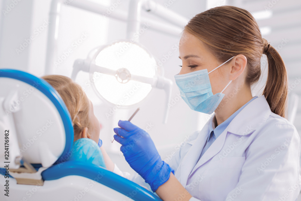 Kind specialist. Enthusiastic reliable dentist feeling satisfied while looking at the condition of teeth of a young patient Kind specialist. Enthusiastic reliable dentist feeling satisfied while looking at the condition of teeth of a young patient