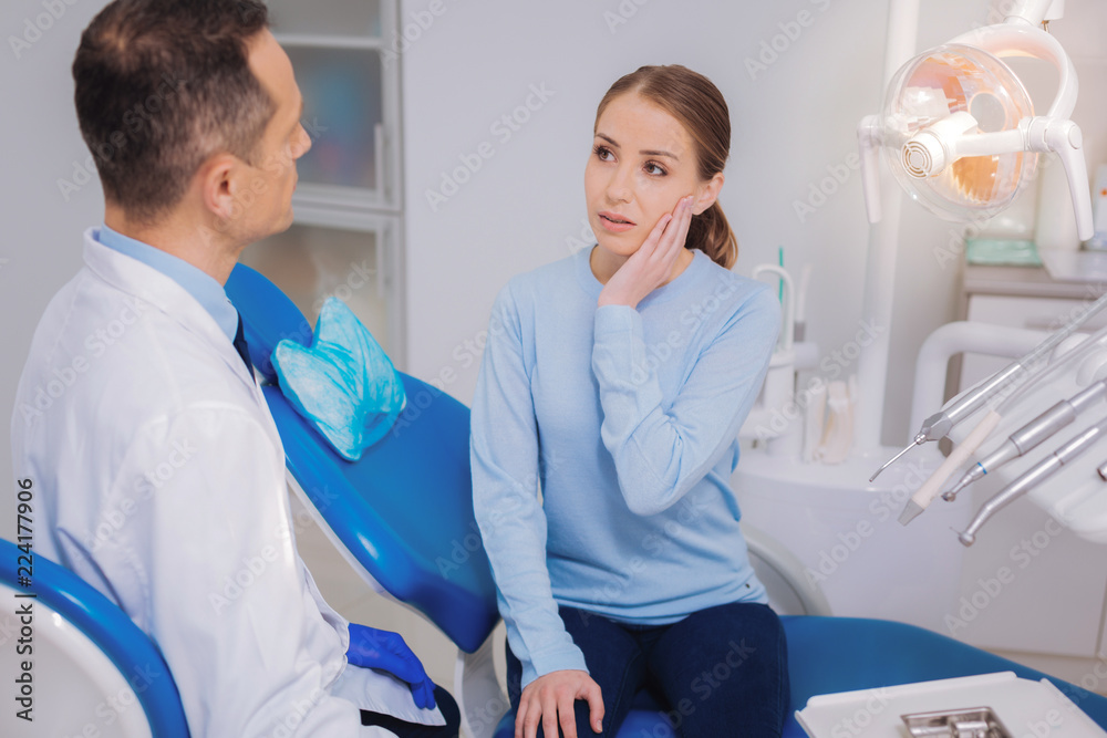 Sharp pain. Tired woman sitting in a dental clinic and touching her cheek while having a toothache