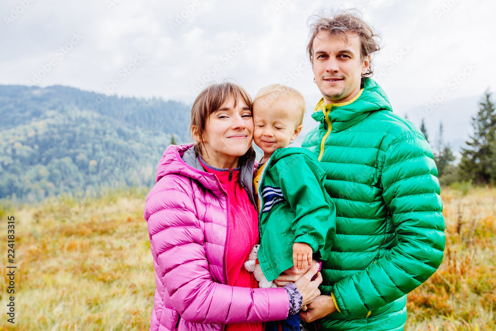 Portrait of happy family mother, father and toddler son wearing colorful duck jacket are traveling on Carpathain mountains. Traveling with children, tourism and people concept. Copy space.
