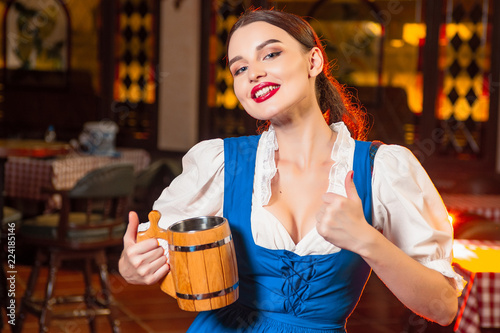 Young beautiful girl with a wooden beer mug in hands at a party Oktoberfest shows thumb up.