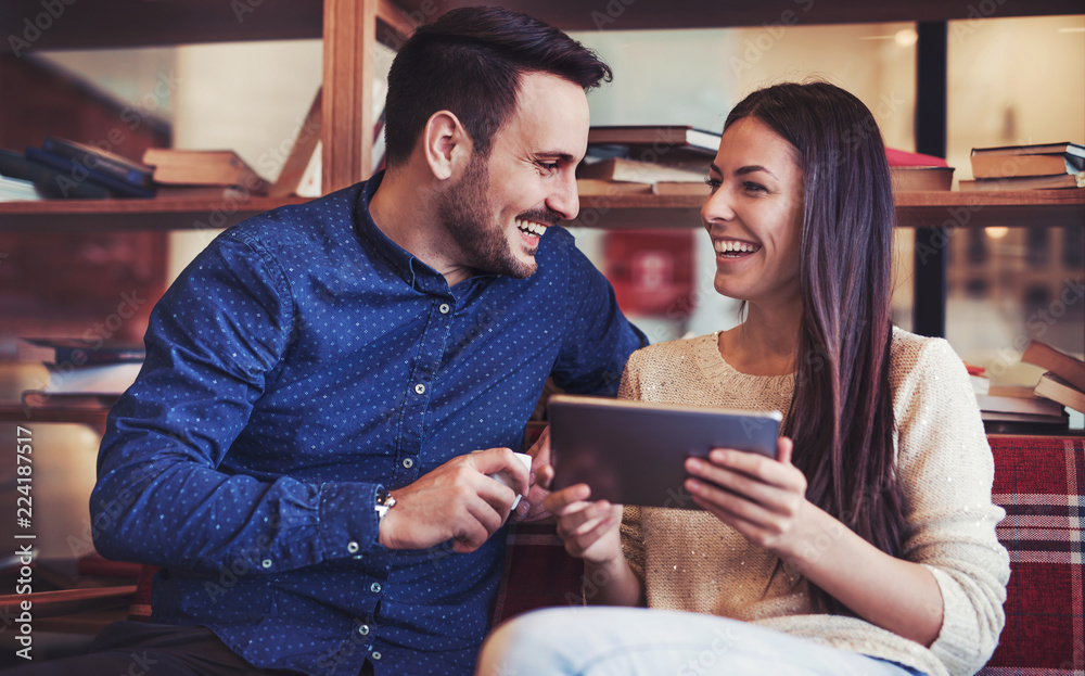 Young couple sitting in the library and having fun with tablet, surrounded with books on the wooden shelves Young couple sitting in the library and having fun with tablet, surrounded with books on the wooden shelves