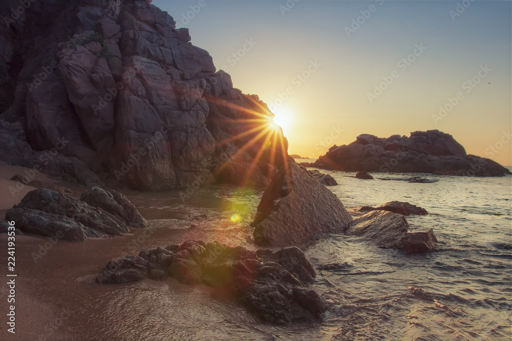 Rocky beach at sunrise with sunrays. Vivid sunbeams behind rock on sea beach. Tropical nature in morning. Paradise lagoon in spain. Rocks and stones on sandy beach Cala sa Boadella in Lloret de Mar.