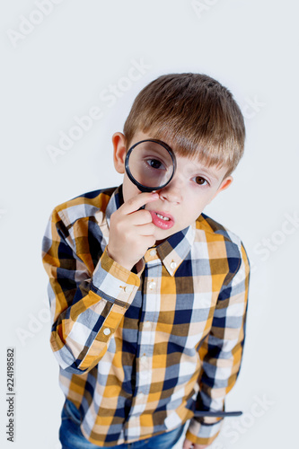 Little boy in a checked shirt on a white background looks with one eye through a magnifier