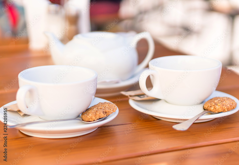 Two white cups of tea and spoons with biscuits, teapot on a wooden table Two white cups of tea and spoons with biscuits, teapot on a wooden table