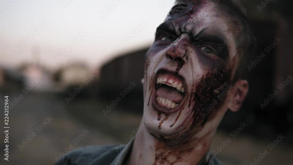 Portrait of a male zombie with bloody teeth and wounded face screaming and shouting. Halloween, filming, staging concept. Blurred railway wagons on the background Portrait of a male zombie with bloody teeth and wounded face screaming and shouting. Halloween, filming, staging concept. Blurred railway wagons on the background