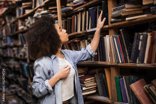 Young female african american librarian making order on bookshelves in college, dark skinned woman student picking literature for education checking information in books store, hipster girl get novels photo