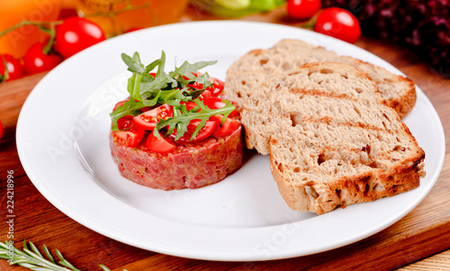 Beef tartare garnished with tomatoes, arugula salad and toasted bread on white plate. Close up
