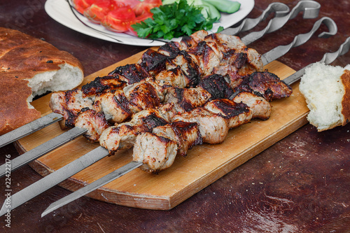 grilled meat on skewers on a wooden tray, bread, vegetables and herbs around, on the table