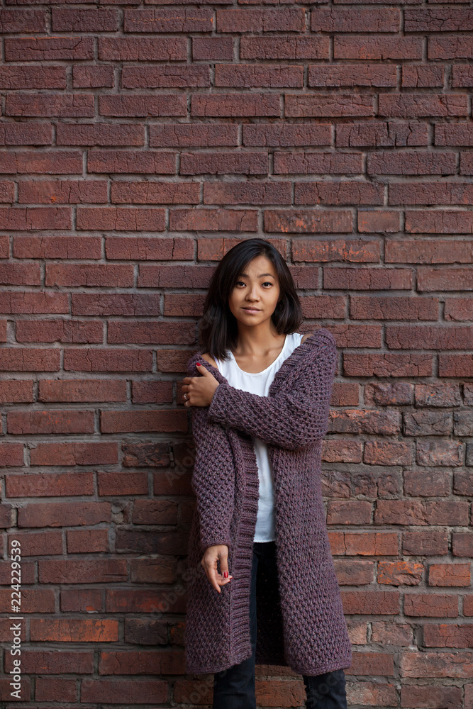 beautiful Asian young woman standing in a knitted purple warm cardigan by a brick wall beautiful Asian young woman standing in a knitted purple warm cardigan by a brick wall