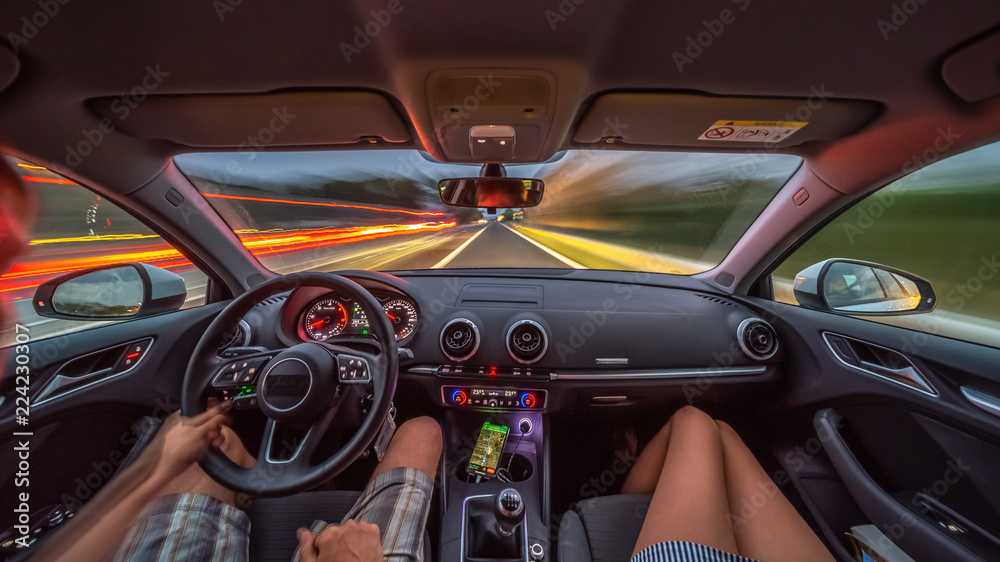 Driver's hands on a steering wheel of a car and woman in the passenger seat. Road trip on the Italians road at evening time.