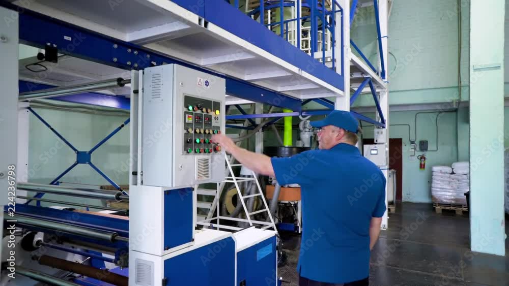 A worker walks at the production site of the plant, inspects, verifies, and adjusts the bag blown film LDPE plastic machine A worker walks at the production site of the plant, inspects, verifies, and adjusts the bag blown film LDPE plastic machine