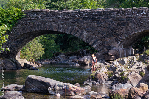 Teenage girl is entering into the River Duddon by old stone bridge in Ulpha in the Lake District National Park, UK. Scenic view of English countryside on a sunny summer day.