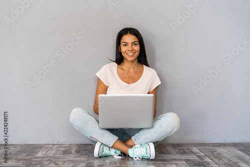 Happy young woman sitting on the floor with crossed legs and using laptop on gray background. photo