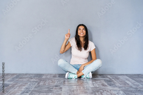 Have idea! Cute woman in casual clothes sitting with legs crossed on the floor gesturing index finger up, meaning eureka over grey wall.