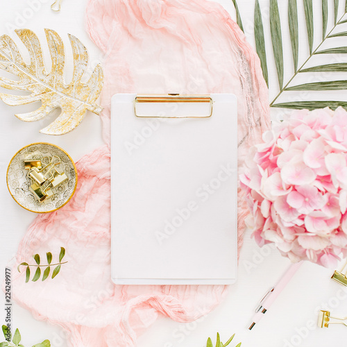 Modern home office desk workspace with blank paper clipboard, pink hydrangea flowers bouquet, tropical palm leaf, pastel blanket, monstera leaf plate and accessories on white background. Flat lay.
