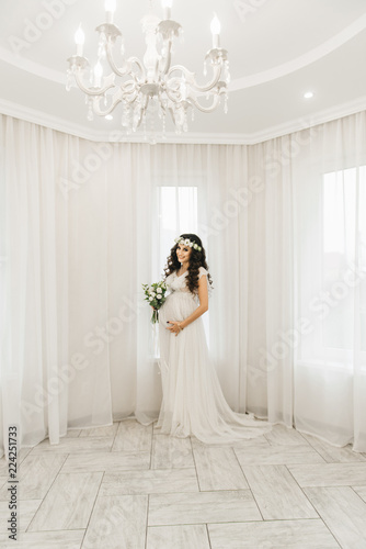 Attractive young pregnant girl in a white dress with curls, a wreath and a bouquet of flowers. Photo session waiting for the baby