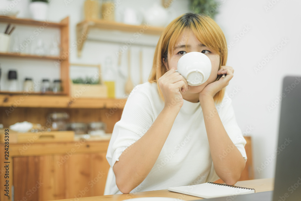 Beautiful young smiling woman working on laptop while enjoying drinking warm coffee sitting in a living room at home. Enjoying time at home. Asian business woman working in her home office. Beautiful young smiling woman working on laptop while enjoying drinking warm coffee sitting in a living room at home. Enjoying time at home. Asian business woman working in her home office.