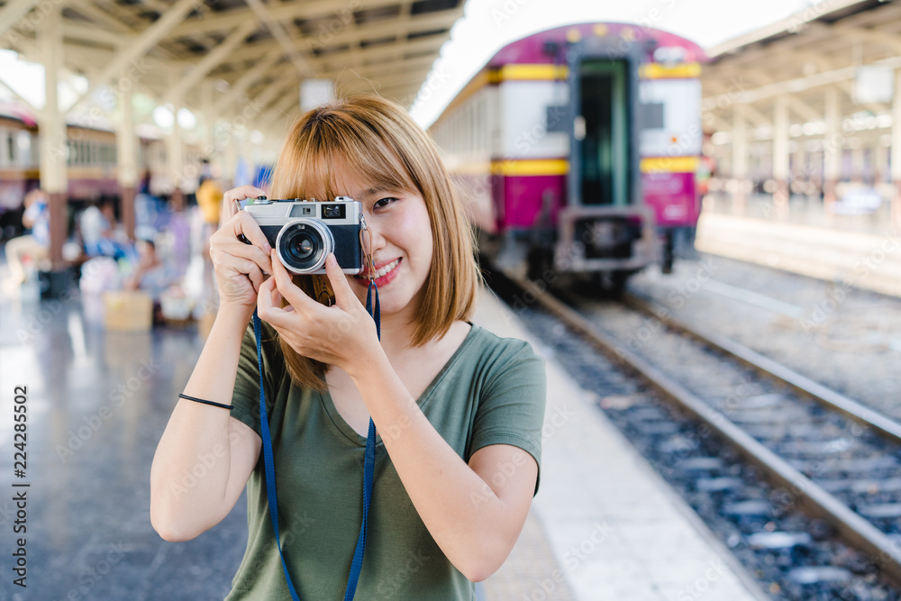 Fototapeta premium Cheerful Traveler backpacker Asian woman using camera to make photo while travel at train station in Bangkok, Thailand. Woman tourist journey on holidays concept.