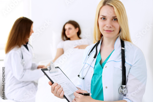 Doctor woman or nurse in a hospital office with her colleague and patient in the background. Healthcare and medicine concept