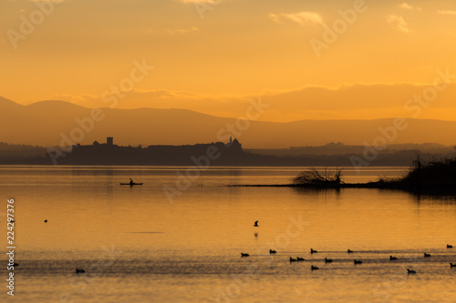 Beautiful view of Trasimeno lake (Umbria, Italy) at sunset, with orange tones, birds on water, a man on a canoe and Castiglione del Lago town on the background