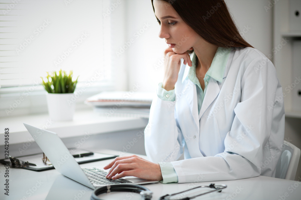 Young female doctor typing on laptop computer while sitting at the table near the window in hospital office Young female doctor typing on laptop computer while sitting at the table near the window in hospital office