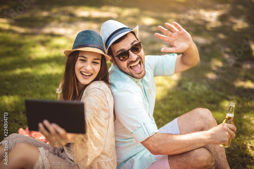 Couple lying on a picnic blanket in a park with a picnic basket filled with fruit, they are using digital tablet