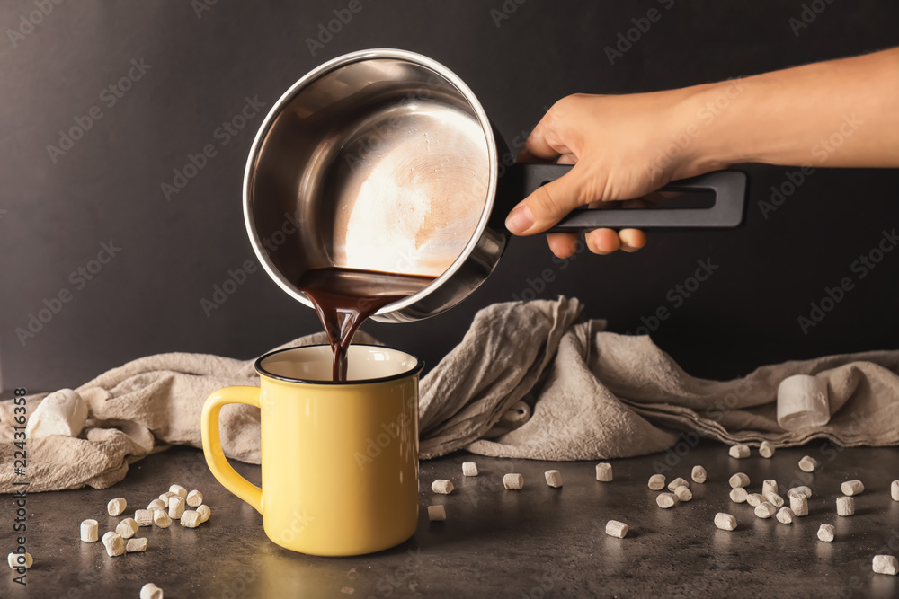 Woman pouring hot chocolate from pot into metal cup on dark table