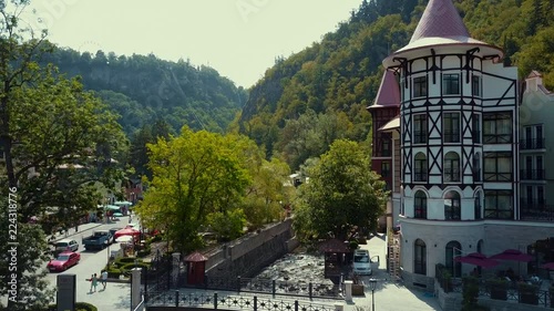 Aerial drone flight up from the river Borjomka close to pine and deciduous trees growing on the sides, Borjomi park in gorge between mountains in sunny day, bridge across the parched river, Georgia
