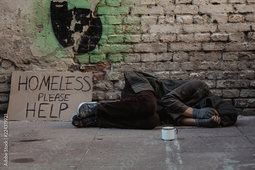 Fotografie Homeless man lies on the street in the shadow of the old building and begging for help and money